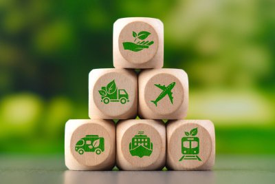 Das Bild zeigt eine Pyramide aus sechs hölzernen Spielwürfeln vor einem unscharfen, grünen Naturhintergrund. Auf den Würfeln sind grüne Symbole abgebildet, die das Thema umweltfreundlicher Transport und nachhaltige Logistik darstellen.